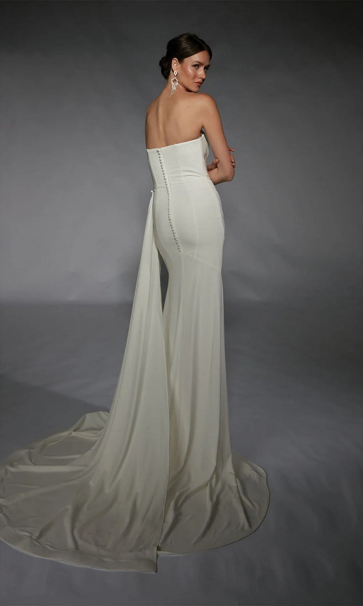 A woman models the Alyce 7105, a strapless long white dress with a train and button details, standing against a gray backdrop and looking over her shoulder.